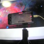 モニターの前に設置された三脚付きのスマートフォン。スマートフォンの画面にはDWARF 3のアプリが表示され、星団のライブ撮影が進行中。背景の大きなモニターには鮮やかなオリオン大星雲のような宇宙の画像が映し出されており、室内からリモートで天体撮影を楽しんでいる様子。