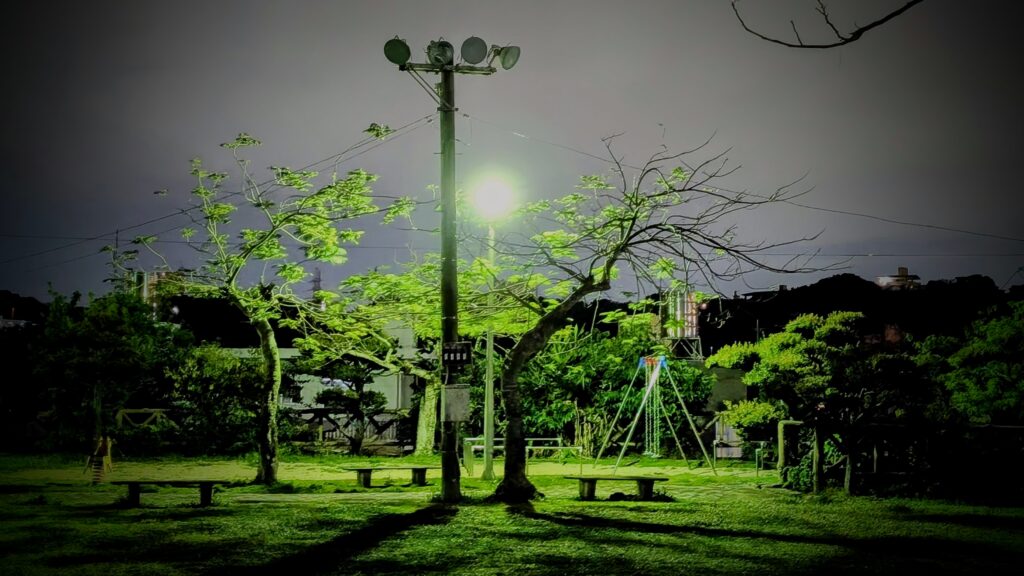 ​この画像は、沖縄の夜、静まり返った無人の公園を描写しています。中央にそびえるユーティリティポールに取り付けられた複数の投光器のうち、中段の強力なスポットライトが、鮮やかな緑色の葉を持つ二本のユーカリのような木と、その下のベンチ、そしてグラウンドを照らし出しています。右側にはブランコセットがたたずみ、背景には夜の街のシルエットと遠くの建物の貯水タンクが見えます。記事で触れられている、夕方から夜にかけての静寂と、冷え込んだ夜の雰囲気を視覚的に強調する一枚です。